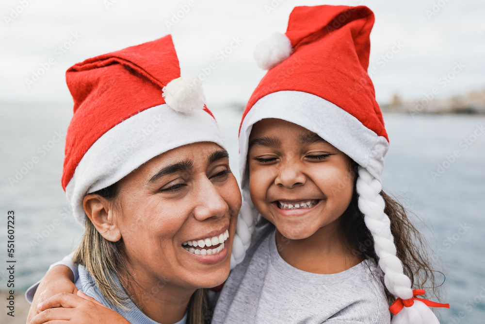 Happy latin mother and little daughter hugging each other outdoor while wearing santa claus hat - Mom and child celebrating christmas together Happy latin mother and little daughter hugging each other outdoor while wearing santa claus hat - Mom and child celebrating christmas together