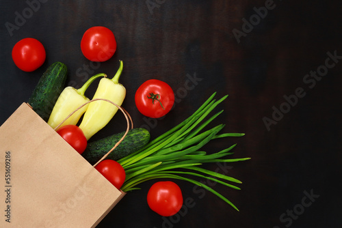 Buying fresh vegetables, vegetarian set. Paper bag with tomatoes, peppers, green onions and cucumbers on a dark table, top view.