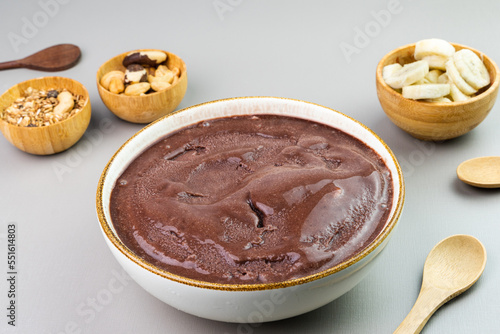 Brazilian açai in a white bowl, additional banana, granola and nuts in small bamboo bowls. Cashew and Pará nuts. Top view. Gray background. Selective focus