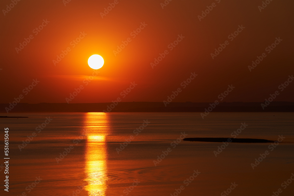Sunset over a flat river. The golden disk of the sun in the red sky is reflected by a golden path in the water, picturesquely bending in small wind waves.