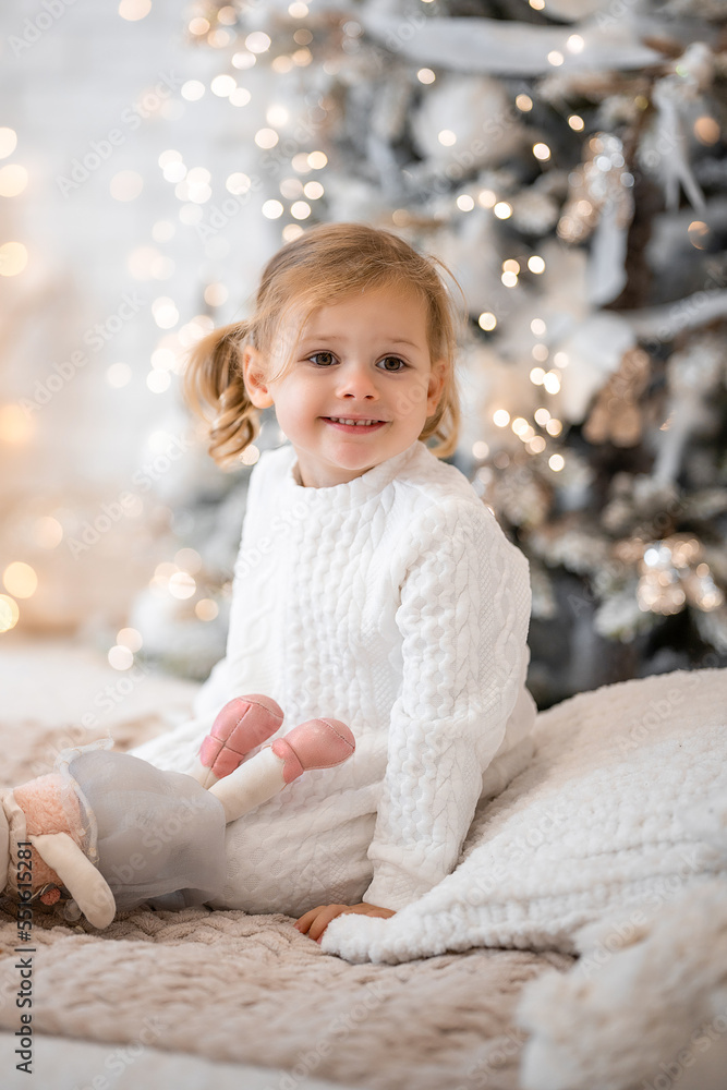 Fototapeta premium Very nice charming little girl blonde in white dress sitting on a child's bed and laughs on the background of Christmas trees in bright interior of the house