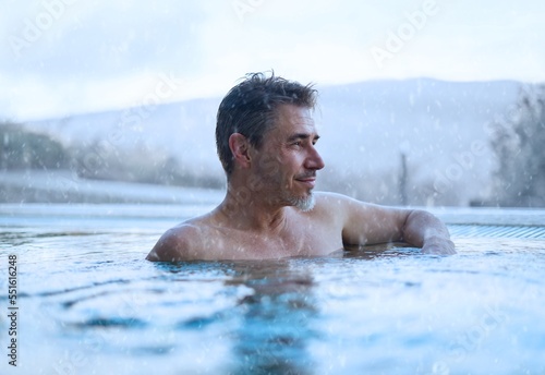 Older handsome man relaxing in outdoor pool on a winter day while it is snowing. Snowy mountains on the background.