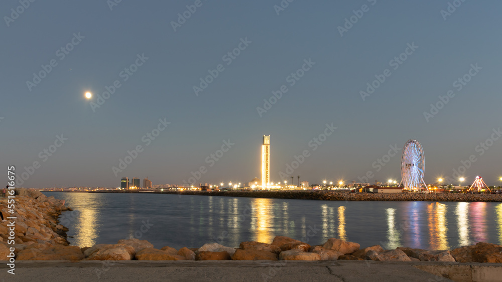 The great mosque and ferris wheel under moonlight seen from Sablette promenade jetty. Skyline view of Algiers city by night, water reflexion lights illuminated by colorful world biggest minaret. The great mosque and ferris wheel under moonlight seen from Sablette promenade jetty. Skyline view of Algiers city by night, water reflexion lights illuminated by colorful world biggest minaret.