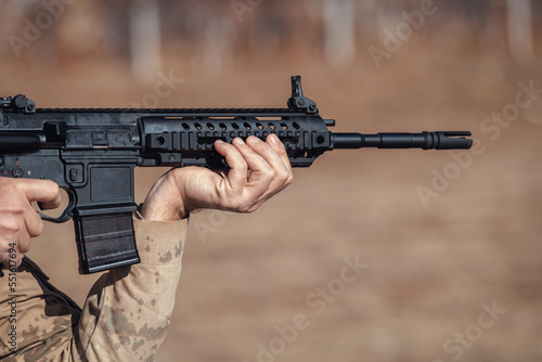 Soldier holding an infantry automatic rifle and aiming. Rifle with modular sight system and cold hammer forging barrel. Firearm shooting and tactical weapons training. Outdoor shooting range.