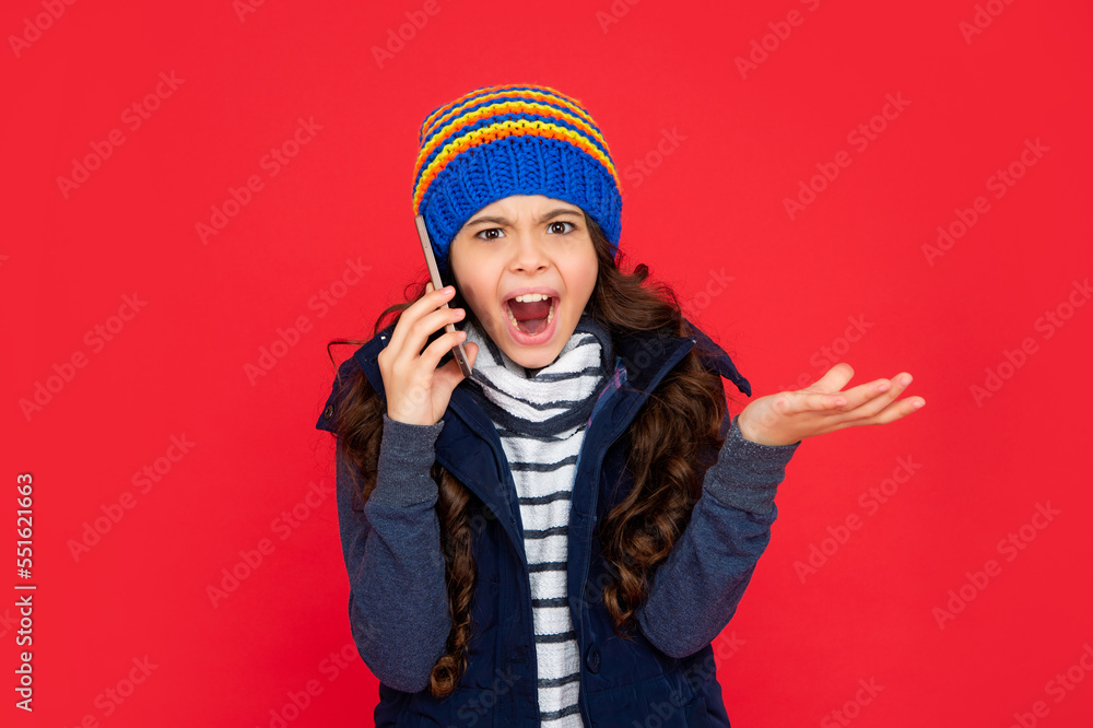 unpleased kid talking on phone in hat on red background. teen girl in down vest with smartphone. unpleased kid talking on phone in hat on red background. teen girl in down vest with smartphone.