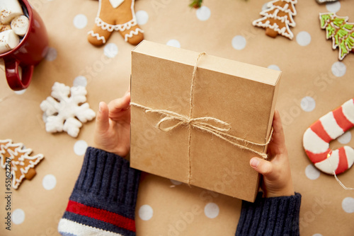 Child holds gift box. Crafting decoration, gingerbread cookies, branch of fir tree, hot beverage with marshmallows and Christmas ornaments. Trendy DIY style. Merry Christmas concept photo