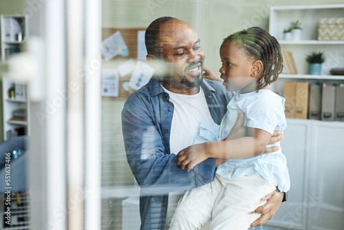 Waist up portrait of happy father embracing cute daughter in office behind glass wall, bring child to work day