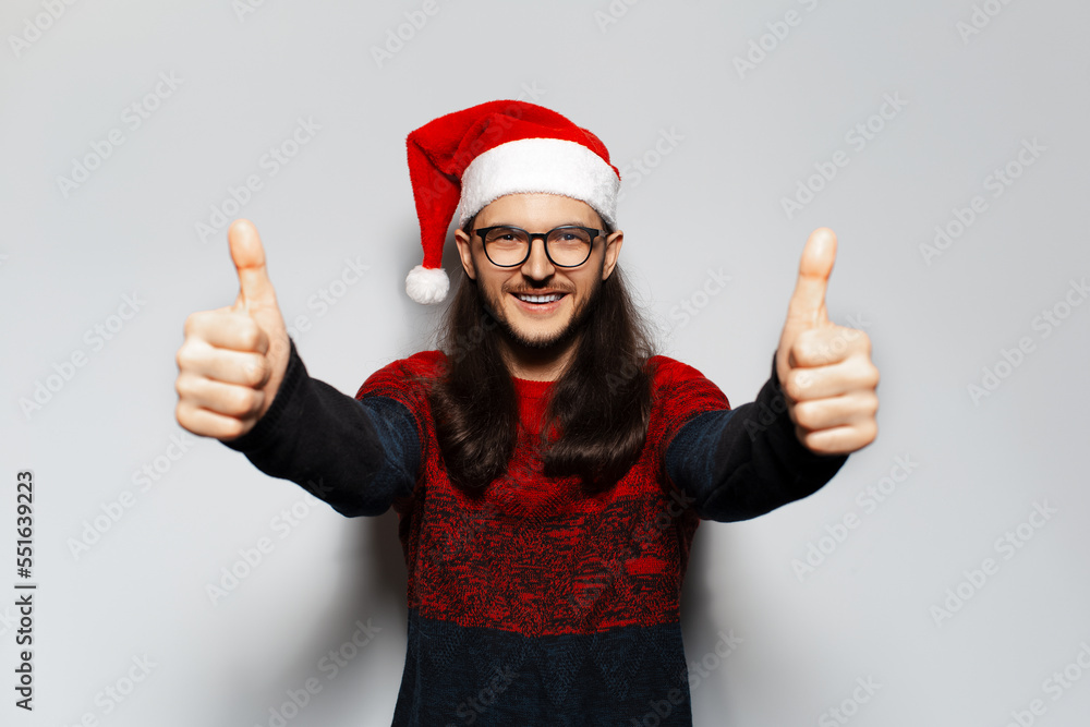 Studio portrait of young happy man with long hair and eyeglasses, showing thumbs up, wears red Christmas sweater and Santa hat on a white background. Studio portrait of young happy man with long hair and eyeglasses, showing thumbs up, wears red Christmas sweater and Santa hat on a white background.