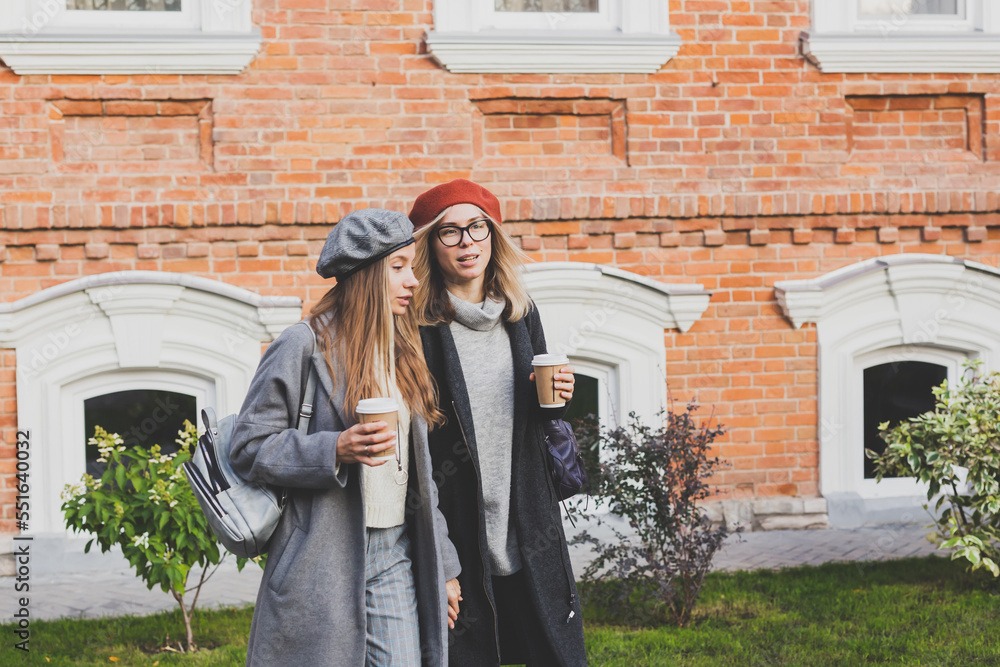 Fototapeta premium Young female best friends talking to each other discussing interesting themes during free time hipster girls joking and having fun together enjoying recreation with coffee to go on urban background