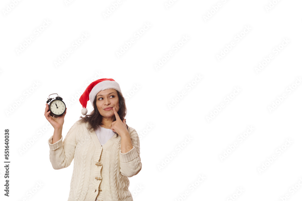 Attractive Latin American woman wearing Santa hat, holding a black alarm clock counting minutes to midnight, holding her finger on lips and dreamily looking aside at copy space. New Year. Christmas Attractive Latin American woman wearing Santa hat, holding a black alarm clock counting minutes to midnight, holding her finger on lips and dreamily looking aside at copy space. New Year. Christmas