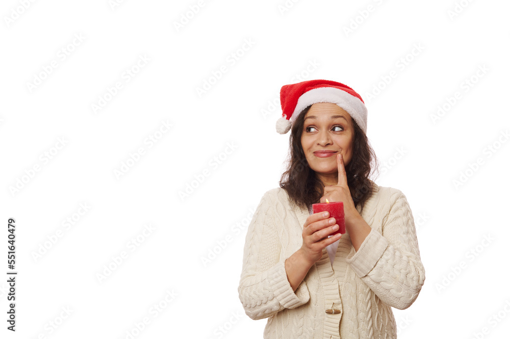 Attractive woman, in Santa hat and beige warm sweater, thoughtfully looks at copy advertising space on white background, puts finger on her lips, posing with a red lit Christmas candle in her hands. Attractive woman, in Santa hat and beige warm sweater, thoughtfully looks at copy advertising space on white background, puts finger on her lips, posing with a red lit Christmas candle in her hands.
