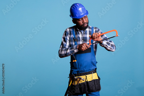 African american carpenter handling hand saw with uniform and protective helmet. Construction worker in coveralls preparing tools isolated on blue background studio shot.