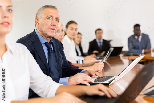 Mature senior white male manager attending business meeting in conference room and intently listening to colleague's report together with coworkers sitting in row