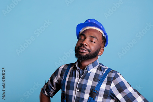 Construction worker with a dream to start work very early in the morning. African american contractor wearing hard hat and work uniform with eyes closed. against blue background, studio shot. © DC Studio