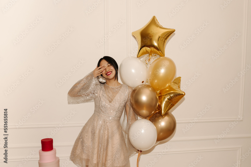 Wonderful young asian girl closing her eyes with hands smiling on white background with balloons. Brunette wears sparkly beige dress on holiday. Concept of enjoying moment