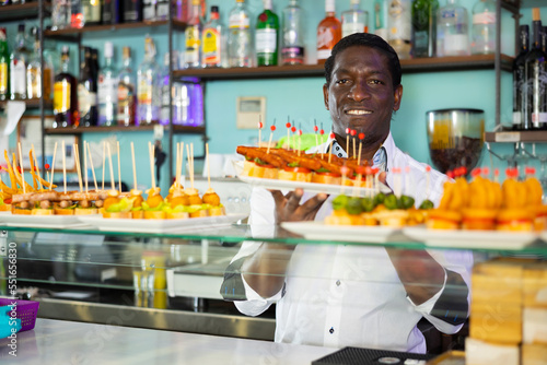 Smiling African American bartender standing with plates of pinchos in front of a pub counter. High quality photo