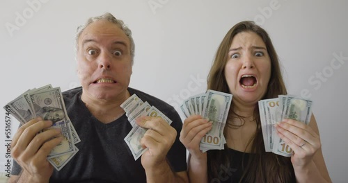 A man and a woman holding and waving stacks of hundred-dollar bills with worried yet comical expressions, looking at the camera as if concerned about their money in a lighthearted and funny way