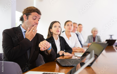 Sleepy businessman looking at his wrist watch and yawning during conference in meeting room.