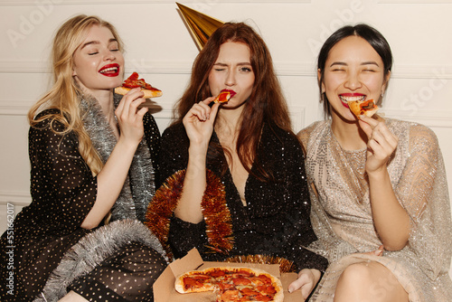 Image of three young multicultural women in sparkly dresses eating pizza sitting on floor. Girlfriends in good mood celebrate birthday at home. Happy weekend concept