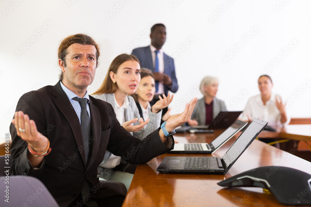 Middle aged white brunette male office worker with questioning facial expression spreading his arms to the sides as if asking what nonsense are you saying, while sitting at corporate team meeting in - obrazy, fototapety, plakaty 