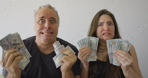 A man and a woman holding stacks of hundred-dollar bills in their hands, with comically worried yet funny expressions, looking directly at the camera, creating a humorous and lighthearted scene
