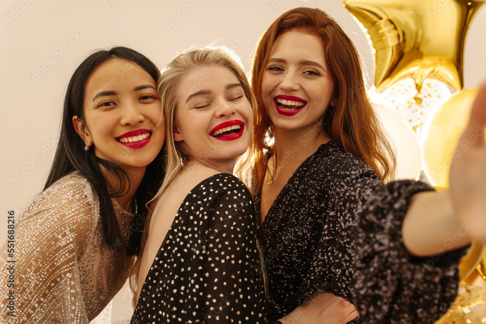 Obraz premium Happy young girls of caucasian and asian appearance take selfie together relaxing on holiday. Pretty women with red lipstick wear trendy dresses. Event concept