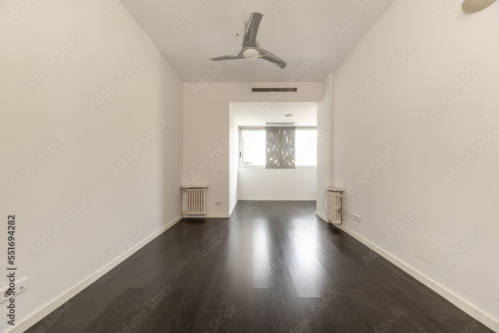 Empty living room of a house with a dark brown laminate floor, several cast iron radiators and a fan with gray blades on the ceiling Empty living room of a house with a dark brown laminate floor, several cast iron radiators and a fan with gray blades on the ceiling