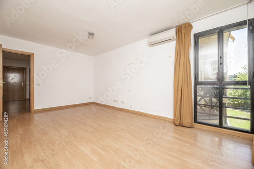 Empty living room of a house with a light brown laminate floor, a bay window with black anodized aluminum windows and light brown curtains