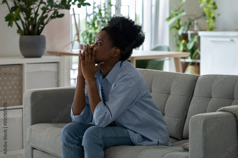 Discouraged pensive African American woman looking into distance thinking about ways to solve financial problems. Frightened black girl sits on sofa in living room having lost mood due to inflation