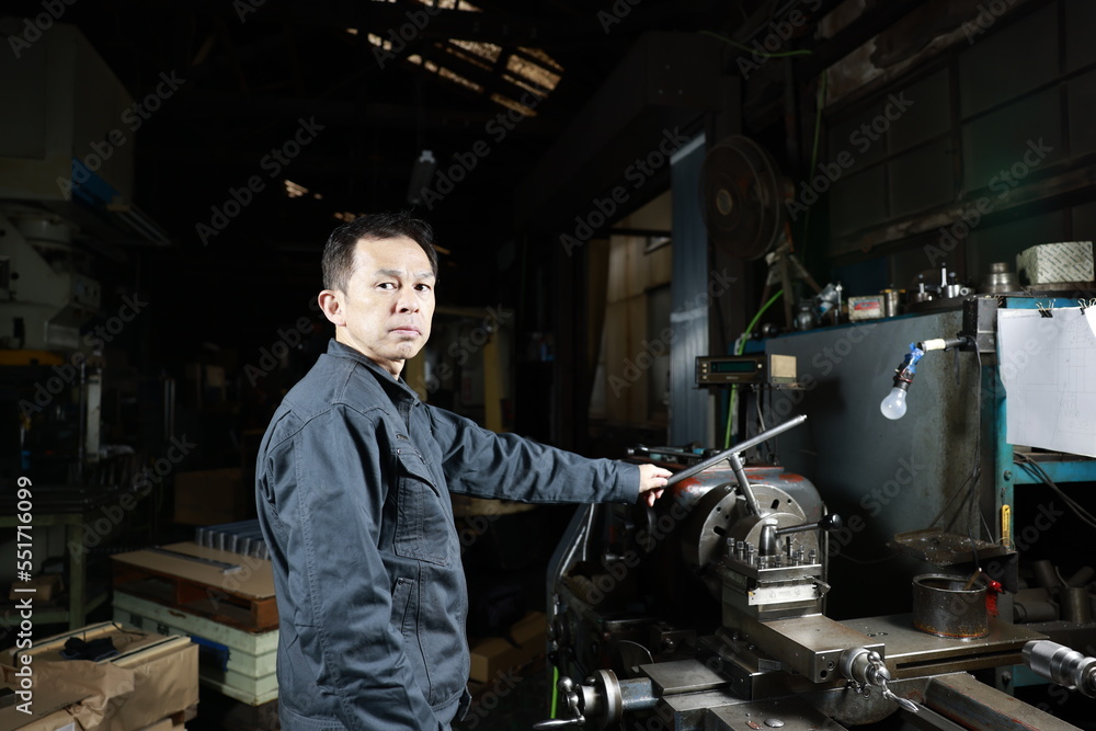 Metalworker in gray work clothes and a lathe in a small to medium-sized small business town factory. Conceptual images of the essence of manufacturing and technical succession. Metalworker in gray work clothes and a lathe in a small to medium-sized small business town factory. Conceptual images of the essence of manufacturing and technical succession.