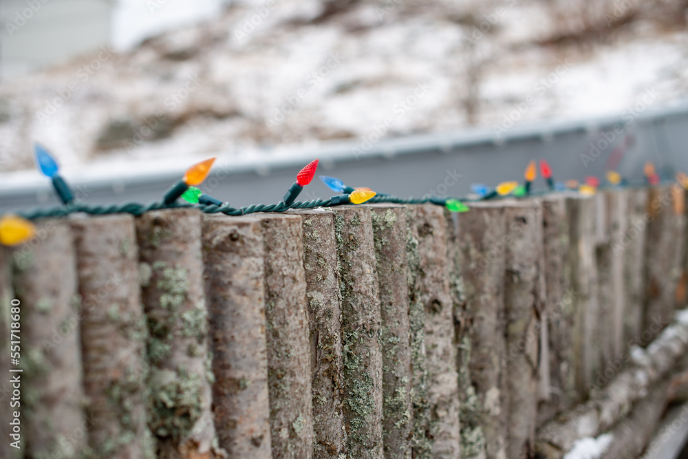 Naklejka premium A fence made with logs, sawed on the ends with bark is on the wood palings. Along the top of the wall is a string of colored Christmas mini-lights. The ocean, snow, and grassy mounds are behind.