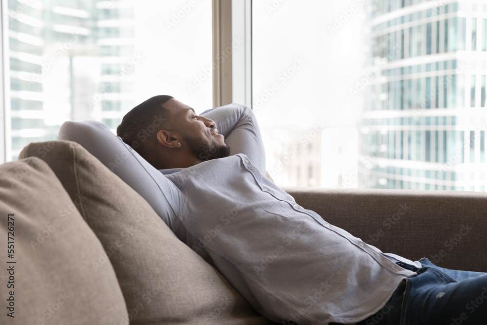 Happy relaxed young African man resting on sofa in home apartment, leaning on back, breathing fresh air, looking at big window, city buildings view, enjoying break, leisure, thinking, dreaming Happy relaxed young African man resting on sofa in home apartment, leaning on back, breathing fresh air, looking at big window, city buildings view, enjoying break, leisure, thinking, dreaming