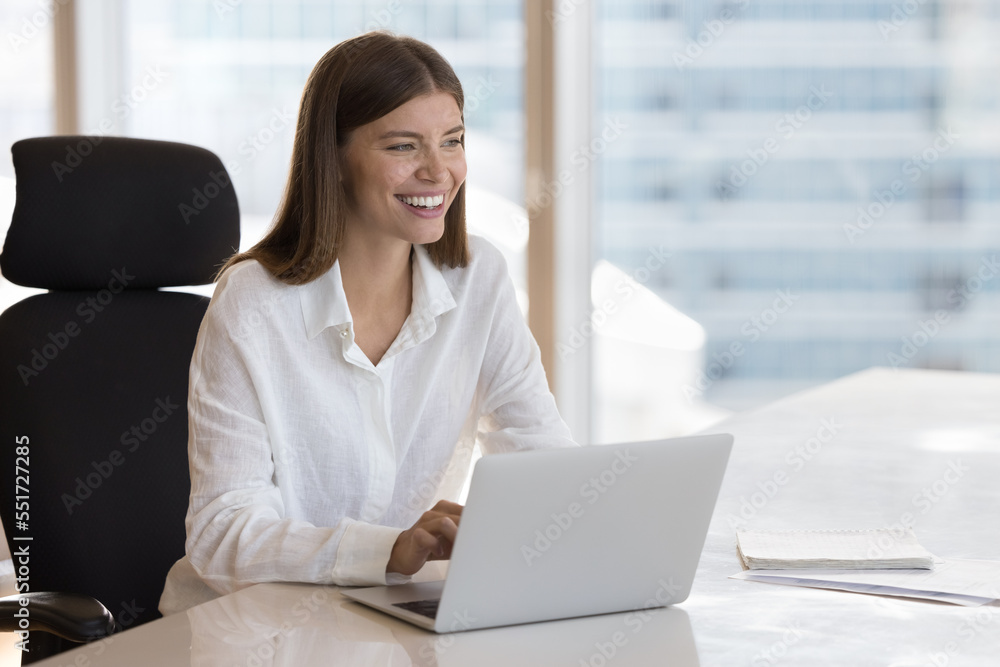 Cheerful happy young office worker sitting at laptop at office work table, typing, looking away, smiling, laughing. Joyful millennial employee, entrepreneur, business woman candid portrait Cheerful happy young office worker sitting at laptop at office work table, typing, looking away, smiling, laughing. Joyful millennial employee, entrepreneur, business woman candid portrait