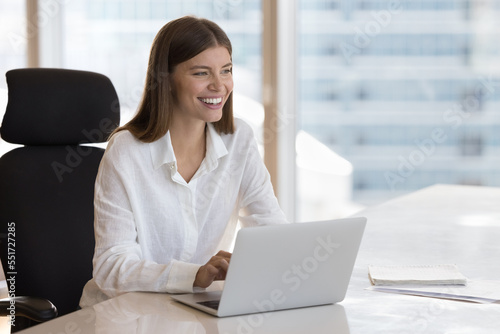 Cheerful happy young office worker sitting at laptop at office work table, typing, looking away, smiling, laughing. Joyful millennial employee, entrepreneur, business woman candid portrait