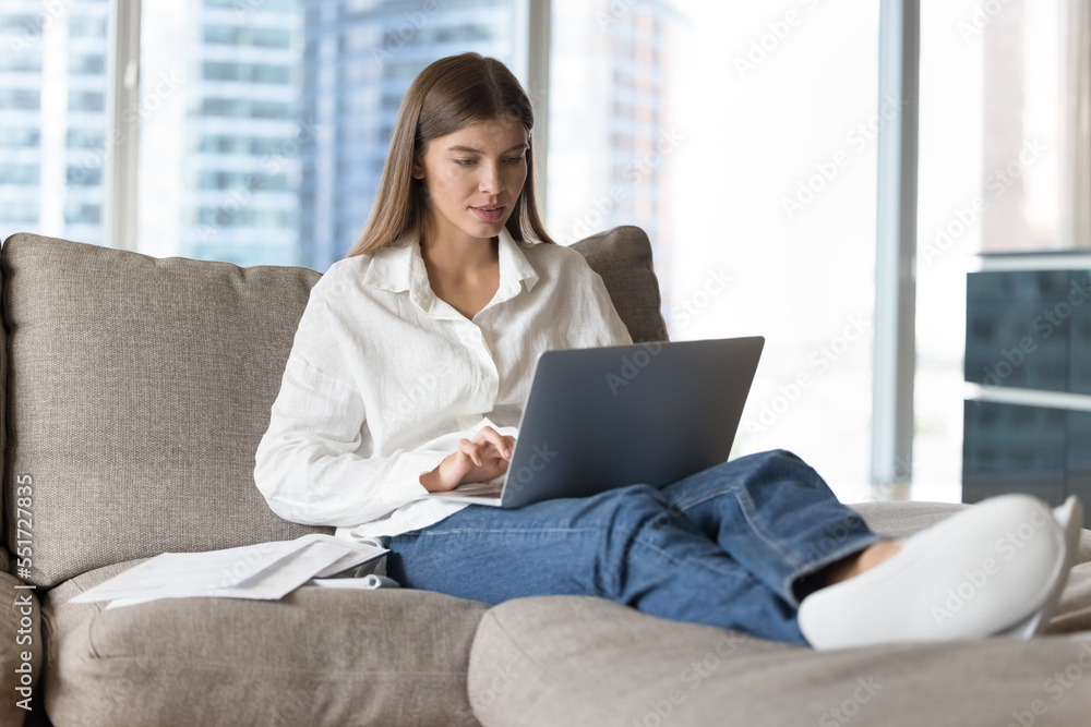 Focused young adult student girl using laptop at home, resting on soft couch at heap of papers, typing. Blogger writing essay, article, chatting, using computer for online communication Focused young adult student girl using laptop at home, resting on soft couch at heap of papers, typing. Blogger writing essay, article, chatting, using computer for online communication
