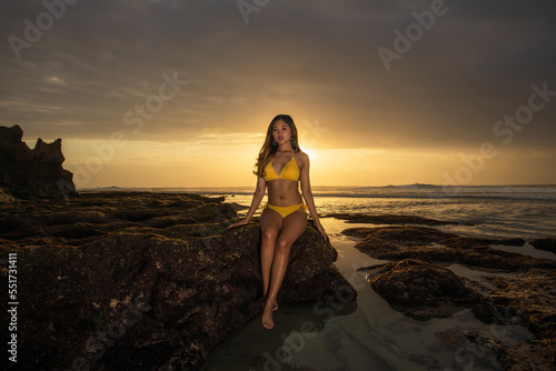 Attractive Asian woman sitting on the rock, wearing yellow bikini. Sexy body. Sunset on the beach. Summer vacation on tropical island. Travel lifestyle. Horizontal layout. Suluban beach Bali
