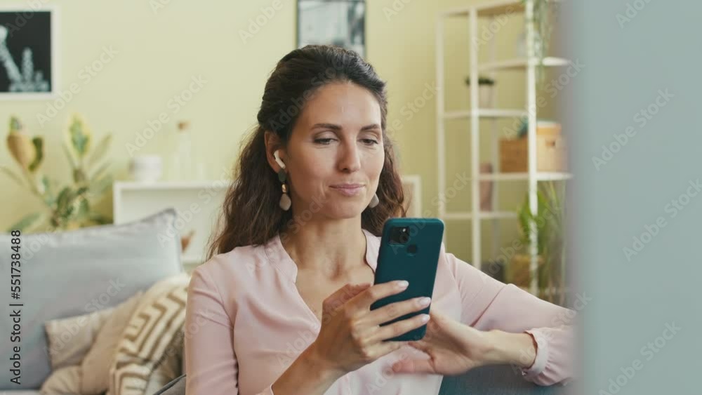 Elegant young Caucasian woman in wireless earbuds using smartphone while talking on video chat with friend, sitting on sofa in living room Elegant young Caucasian woman in wireless earbuds using smartphone while talking on video chat with friend, sitting on sofa in living room
