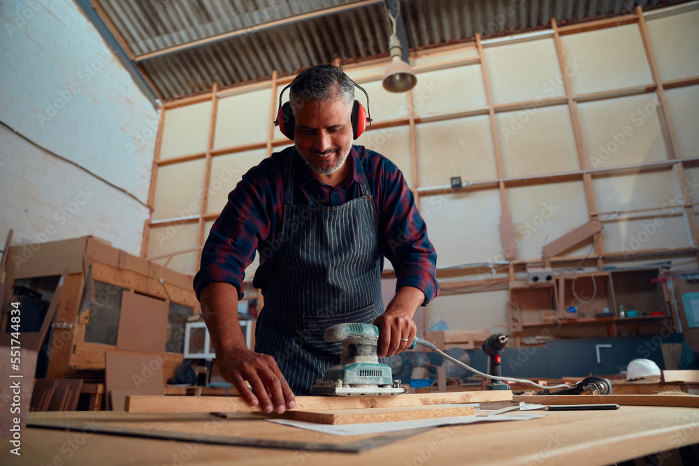 Mid adult man smiling while wearing ear muffs and using power tool on timber in woodworking factory Mid adult man smiling while wearing ear muffs and using power tool on timber in woodworking factory