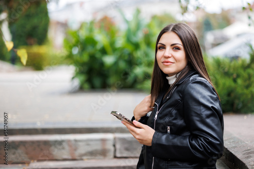 A young pretty girl wearing a black leather jacket walks down the street in a park and uses her smartphone for work. The work of a freelancer and blogger.