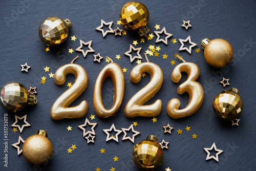 The golden figures 2023 made of candles on a black stone slate background are decorated with a festive decor of stars, sequins, fir branches, balls and garlands. Greeting card, happy New Year.