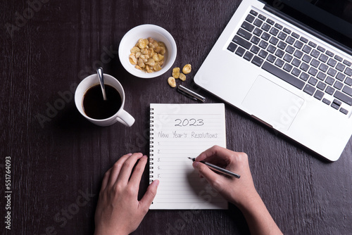 Conceptual,Top view office desk mockup: laptop, notebook, smartphone, snack bean, and cup of coffee on rustic brown wooden desk with hand writing. New year 2023 resolution