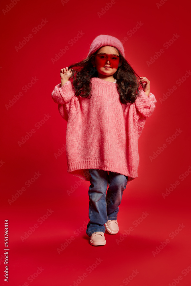 Portrait of stylish little girl, child, posing in oversized pink sweater, red glasses and knitted hat isolated over red background. Concept of childhood, fashion, emotions Portrait of stylish little girl, child, posing in oversized pink sweater, red glasses and knitted hat isolated over red background. Concept of childhood, fashion, emotions