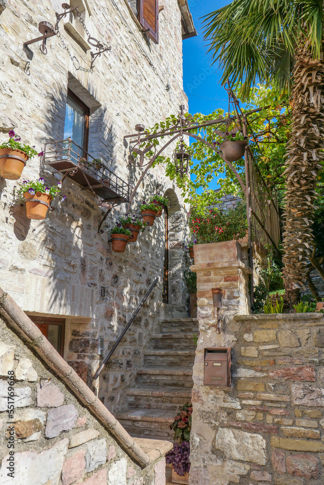 A stone Italian house and a staircase to the top. There is a small ivy-covered canopy above the stairs, and pots of flowers hang on the left wall of the house.