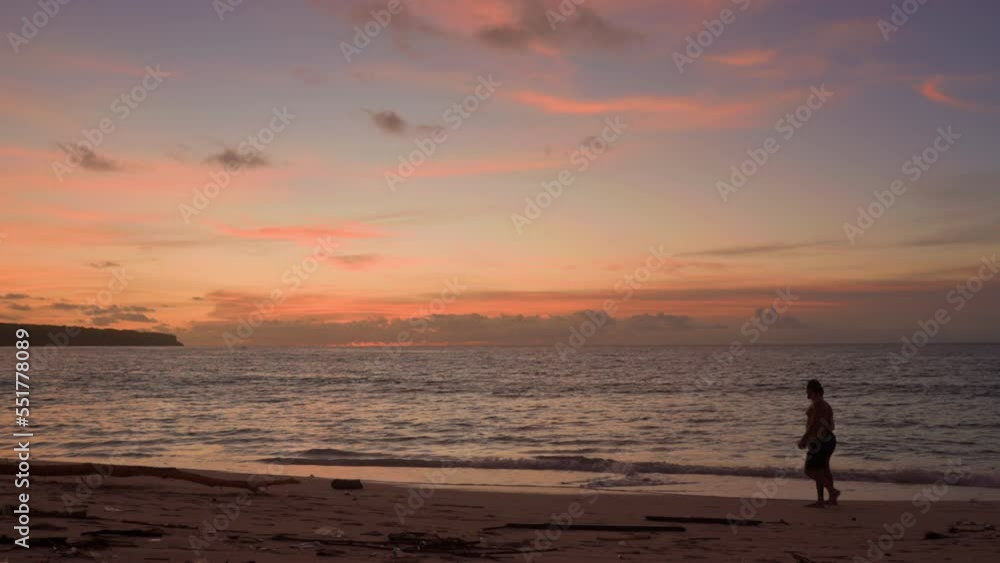 Silhouettes of two men in beach shorts strolling along beach in ocean during sunset. Tourists walk on sand against background of the sea and colored sky of warm orange tones. Relaxing holiday on shore Silhouettes of two men in beach shorts strolling along beach in ocean during sunset. Tourists walk on sand against background of the sea and colored sky of warm orange tones. Relaxing holiday on shore