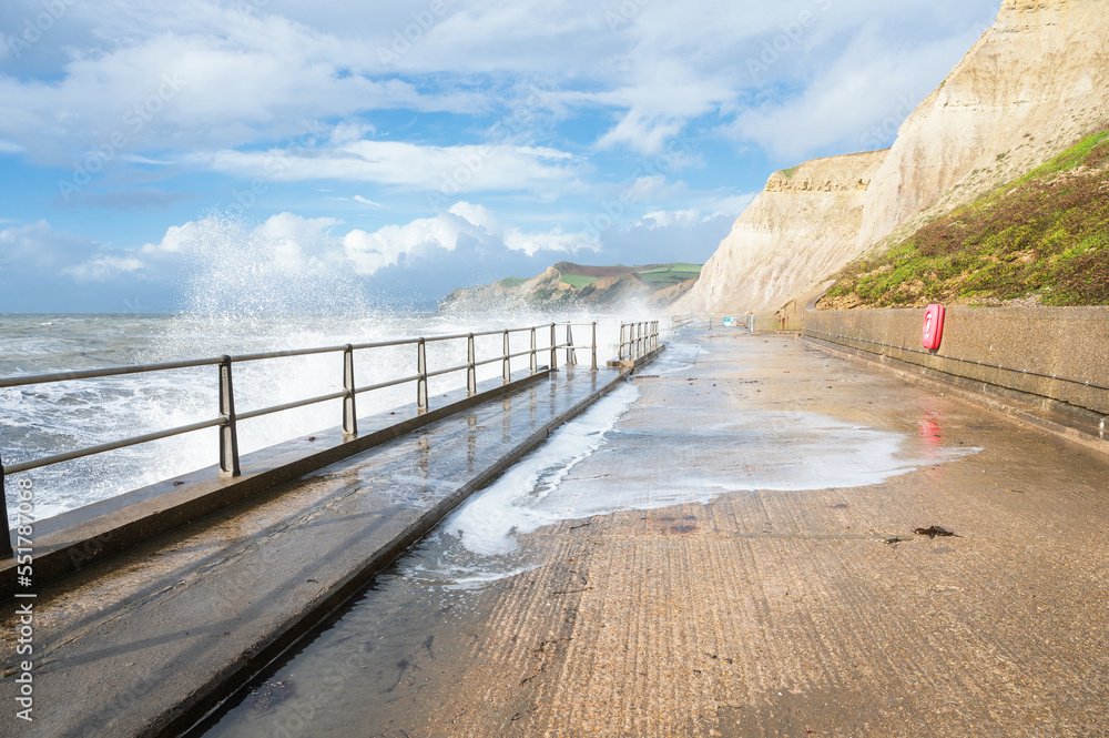 Big waves and water splashes at West Bay beach near Bridgport in Dorset, United Kingdom. Walkway or promenade along the beach, selective focus