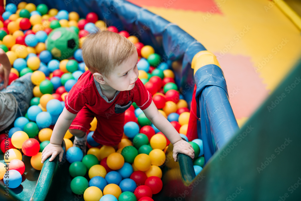 Obraz premium Happy laughing boy 1-2 years old having fun in ball pit in kids amusement park and indoor play center. Activity toys for little kid