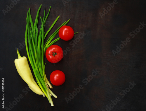 Vegetables lie on a dark brown wooden table, top view. Green onions, yellow peppers and ripe red tomatoes, healthy eating concept