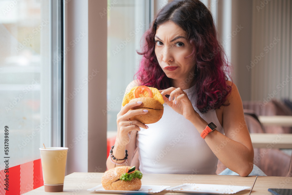 Frowned disgruntled girl customer of a fast food restaurant digs into a burger and found a spoiled product in her lunch Frowned disgruntled girl customer of a fast food restaurant digs into a burger and found a spoiled product in her lunch
