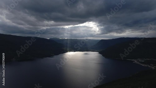 Aerial shot of lake in between the mountains with sunrays coming out from the clouds. Dramatic dark clouds and sun rays over the  valley during the summer. 
 photo