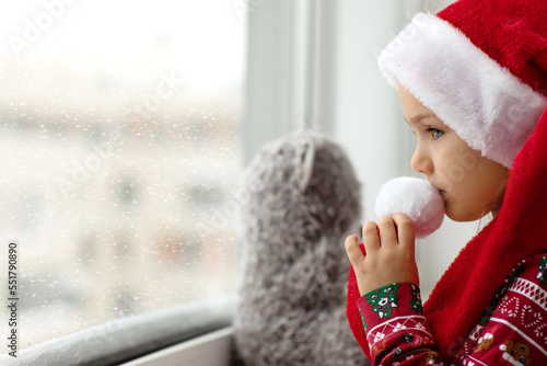 Sad alone child girl in Christmas red Santa hat looks at window and waiting holidays with teddy bear toy. Mental health